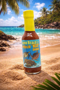 Hot sauce bottle with a yellow cap on a sandy beach with ocean and palm trees in the background